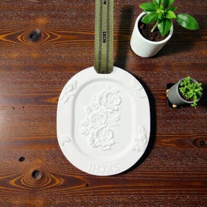 Könnte beinhalten: Weiße ovale dekorative Platte mit erhabenem Blumendesign, aufgehängt an einem grünen Band. Die Platte zeigt ein detailliertes Relief von Rosen. Das Wort "SXCHI" steht unten. Zwei kleine Topfpflanzen sind im Hintergrund.