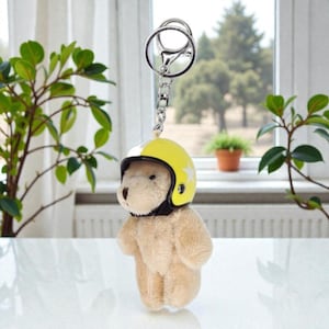 May include: A plush teddy bear keychain wearing a yellow and white motorcycle helmet with a star design. The keychain has a silver metal ring and chain. The bear is light brown and has a black helmet strap.