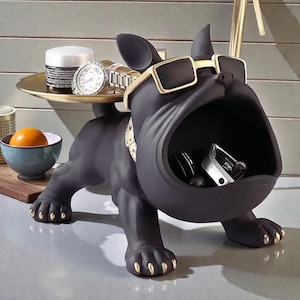 May include: A black bulldog ornament with gold detailing, wearing sunglasses and a collar. The dog's open mouth functions as storage. A gold tray on its back holds a watch and a jar. The dog's paws have gold-painted nails.