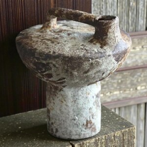 May include: A handcrafted ceramic jug with a handle. The jug has a bulbous body and a cylindrical base. The color is a mix of brown, white, and gray, with a textured, aged appearance. The jug is displayed on a wooden surface.