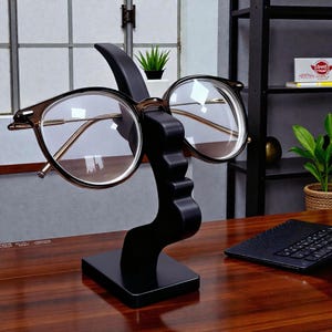 Peut inclure: Un présentoir à lunettes noir avec un design incurvé unique, tenant une paire de lunettes à monture ronde. Le présentoir est sur un bureau en bois, avec un clavier et une fenêtre en arrière-plan.