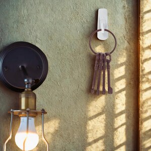 May include: A white adhesive hook holds a key ring with several antique keys. The keys are a dark brown color, and the key ring is a similar shade. The hook is mounted on a textured, light-yellow wall.