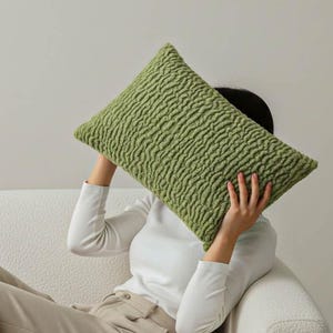 May include: A rectangular, textured, olive green pillow. The pillow is held up in front of a person's face. The person is wearing a white long-sleeve shirt and tan pants, sitting on a white couch.