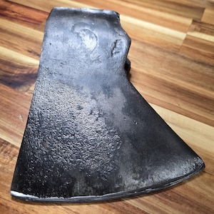 May include: A dark gray metal axe head with a textured, weathered surface and a wide, curved cutting edge. The top portion features a flat poll, while the body shows signs of oxidation and wear, displaying a rugged, industrial appearance.