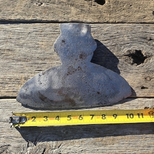 May include: A vintage, weathered metal axe head with a curved blade, measuring approximately 11 inches in length. The axe head is placed on a rustic wooden surface, with a yellow measuring tape alongside it.