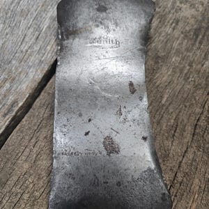 May include: Close-up of a vintage axe head with a weathered, metallic finish. The axe head has a curved blade and a flat top, with the word "Zenith" stamped on the surface. The axe head is resting on a wooden surface.