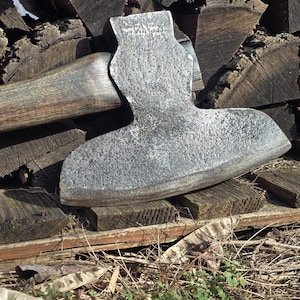 May include: A weathered, gray axe with a wooden handle rests on a pile of firewood. The axe head is textured and shows signs of use. The wooden handle is a light brown color. The axe is positioned horizontally, suggesting it is ready for use.
