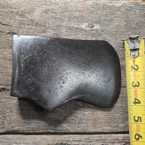 May include: A vintage, dark gray axe head with a curved blade. The axe head is positioned on a weathered wooden surface. A yellow measuring tape is next to the axe head, showing measurements in inches.