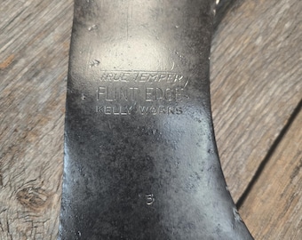 Puede incluir: Primer plano de una cabeza de hacha de doble filo vintage. La cabeza del hacha es de color gris oscuro con desgaste visible. El texto "TRUE TEMPER, FLINT EDGE, KELLY WORKS" está estampado en el metal. También es visible el número "3".