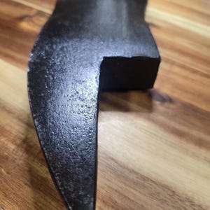 May include: A dark, textured metal tool component with a curved, pointed blade section and a thick, rectangular block-shaped base. The surface of the object appears rough and matte, showing signs of wear and a dark, oxidized finish throughout.
