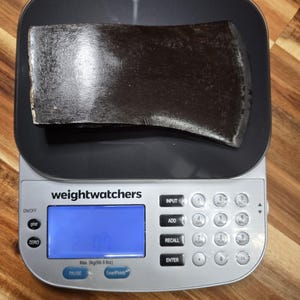 May include: A dark brown axe head rests on a black digital scale. The scale, branded "weightwatchers", displays a blue screen with numbers. The axe is positioned centrally, showing its blade and overall shape.