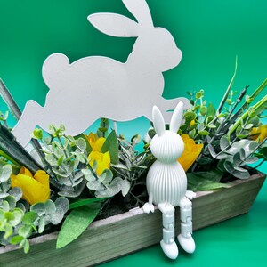 May include: A white, leaping bunny silhouette and a seated bunny figurine are featured in a wooden planter box filled with yellow tulips and greenery. The background is a solid teal color, creating a spring-themed display.