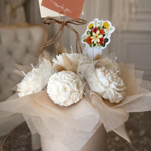 May include: A cream-colored floral arrangement featuring rose-shaped candles, tulle, and dried grasses. A small card with "thank you" is attached. A sticker with a floral design is also included.