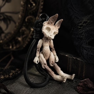 May include: A small, articulated figurine with a skull-like head and animal-like ears. The figure is primarily beige and brown, with a black cord attached for hanging. The doll is posed in a seated position, with its legs extended.