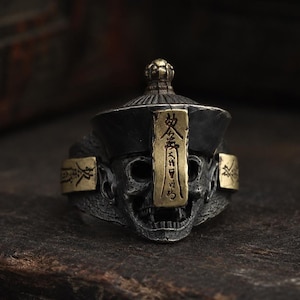 Op de afbeelding: Een zilverkleurige metalen ring met een schedel die een hoed in traditionele stijl draagt. Het ontwerp bevat een verticaal messingkleurig plaatje op het voorhoofd met gegraveerde tekens en bijpassende messingkleurige zijaccenten met inscripties.