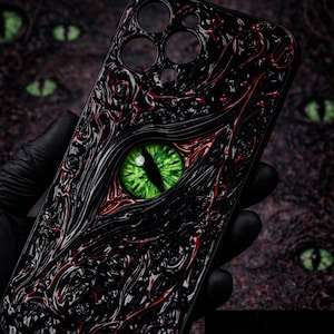 Op de afbeelding: Zwarte telefoonhoes met een gedetailleerd ontwerp met een gloeiend groen oog. De hoes heeft een getextureerd oppervlak met rode accenten. Het oog gloeit in het donker en voegt een uniek visueel element toe.