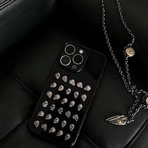 Puede incluir: Funda de teléfono negra con un patrón de pequeñas calaveras blancas. Junto a ella hay un collar de cadena plateada con un colgante decorativo. El fondo es una superficie de cuero negro.