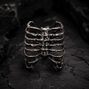 May include: A silver-colored ring shaped like a human rib cage. The ring has a detailed, three-dimensional design with visible ribs and sternum. The ring is set against a dark, textured background.