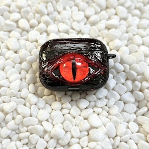 Könnte beinhalten: Schwarzes AirPod-Etui mit einem auffälligen roten Augendesign. Das Auge hat eine schwarze Pupille und ein strukturiertes, reptilienartiges Aussehen. Das Etui befindet sich vor einem Hintergrund aus kleinen, weißen Kieselsteinen.
