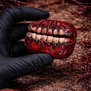 May include: A gruesome, realistic prop of a set of teeth, with a red, fleshy exterior and off-white teeth. The prop is held by a gloved hand, with a blurred background of similar props and a textured surface.