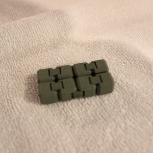 May include: A small, olive green fidget toy composed of eight interlocking blocks. The blocks are arranged in a rectangular shape and sit on a white, textured surface. The toy appears to be made of a solid material.