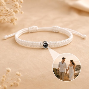 May include: A white woven cord bracelet featuring a central silver-toned circular charm. The charm contains a small, circular projection lens that displays a photograph of a couple standing together outdoors near a body of water at sunset.