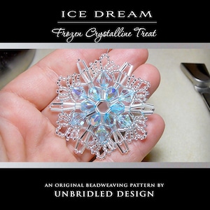 Può includere: Un fiocco di neve fatto a mano, con perline, con un gruppo centrale di cristalli azzurri e perline trasparenti. Il fiocco di neve è tenuto in una mano, con il testo "ICE DREAM Frozen Crystalline Treat" e "AN ORIGINAL BEADWEAVING PATTERN BY UNBRIDLED DESIGN".