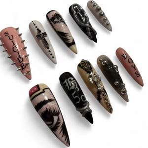Puede incluir: Una colección de uñas artificiales largas y puntiagudas con varios diseños. Algunas uñas son rosas con púas plateadas y la palabra "SUICIDE". Otras presentan arte de ojos, cruces y la palabra "BOYS". Las uñas son en tonos rosa, blanco, negro y plata.