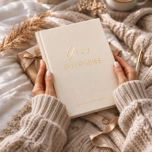 May include: A cream-coloured hardcover book with gold foil lettering that reads Your INTERVIEWS and BABY'S MEMORIES. The book is held against a soft, textured beige fabric background with a wrapped gift and a gold pen visible nearby.