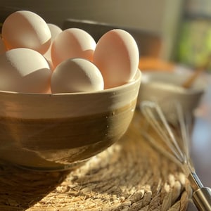 Puede incluir: Un cuenco de cerámica lleno de huevos blancos frescos. El cuenco tiene una combinación de colores marrón claro y beige. Los huevos están enfocados, con un batidor y otros cuencos ligeramente borrosos en el fondo. El cuenco está sobre un salvamanteles tejido.