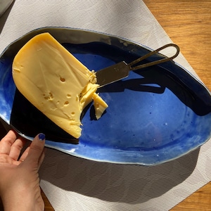 Puede incluir: Una porción de queso amarillo con agujeros está sobre una fuente de cerámica azul. Un pequeño cuchillo para queso descansa sobre el queso. La fuente tiene un borde azul oscuro y un interior azul claro.