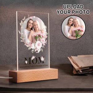 May include: A personalized acrylic photo display with a wooden base. The display features a photo of two women surrounded by a floral wreath. The word "MOM" and the phrase "everything I am is because of you" are printed on the display.
