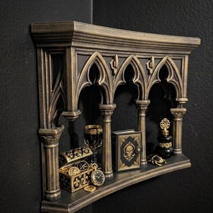 May include: A gothic-style shelf in bronze and black, adorned with miniature treasures. It features arched openings and holds a goblet, a book with a skull, a treasure chest, a pocket watch, and a perfume bottle, creating a dark aesthetic.