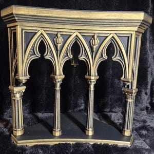 May include: A corner shelf featuring a black base and a gold-toned frame with architectural details. The structure includes four vertical columns supporting pointed gothic-style arches and a flat top surface with a decorative molded edge.