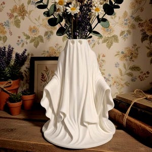 May include: A white ceramic ghost-shaped vase holding a bouquet of white daisies, purple lavender, and green eucalyptus leaves. The vase has a draped fabric design with eye-shaped openings. The vase is on a wooden shelf.