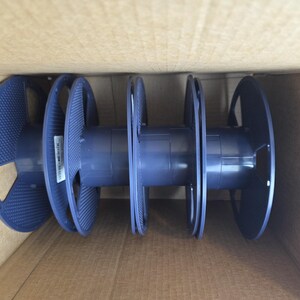 May include: A set of three dark blue plastic spools connected by a central cylindrical core. Each spool features a textured circular side panel with cutouts. The spools are positioned horizontally inside a plain cardboard box container.
