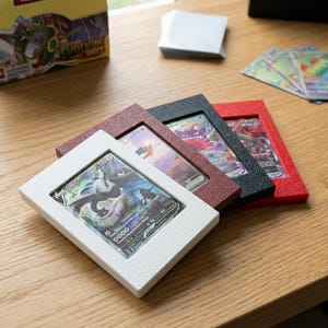 May include: Four card display frames in white, burgundy, black, and red. Each frame holds a trading card with a colorful illustration. The frames are on a wooden surface, with a box of cards and a stack of card sleeves in the background.