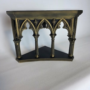 May include: A corner shelf featuring a black triangular base and a decorative front panel with four vertical columns supporting pointed gothic arches. The structure is finished in a metallic antique gold tone with black accents in the recessed areas.