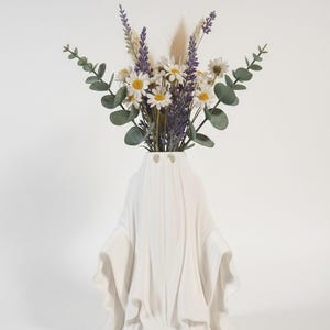 May include: A white ceramic ghost-shaped vase holding a bouquet of flowers. The bouquet includes white daisies, purple lavender, and green eucalyptus leaves. The vase has a draped design, with two eye-shaped openings.