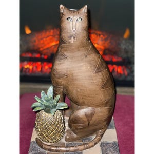 May include: A brown cat figurine with a pineapple. The cat is seated with a detailed, textured design. The pineapple is gold and green. The figurine sits on a black and white checkered base.