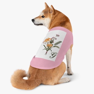 Puede incluir: Un perro Shiba Inu con una camiseta rosa claro. La camiseta presenta un rectángulo blanco con un ramo floral y la letra "G" en cursiva. El ramo incluye flores naranjas y moradas.