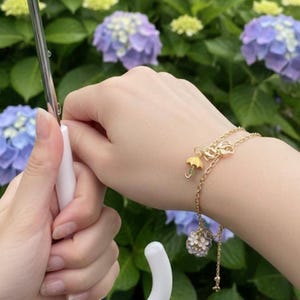 May include: A gold bracelet featuring two chains and charms. One charm is a small umbrella, and the other is a cluster of white flowers. The bracelet is worn on a wrist, with a white umbrella handle in the background.