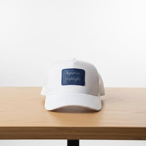May include: White trucker hat with a mesh back and a curved brim. The hat features a blue rectangular patch on the front with the words "Aquarius Highlights" in white lettering. The hat is sitting on a wooden surface.