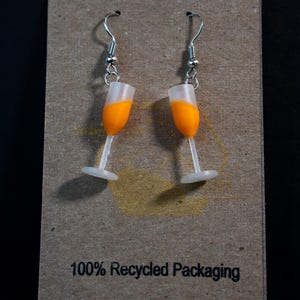 May include: A pair of champagne glass earrings with orange liquid inside. The earrings are made of white and orange plastic and hang from silver hooks. The earrings are displayed on a brown cardboard backing with the text "100% Recycled Packaging".