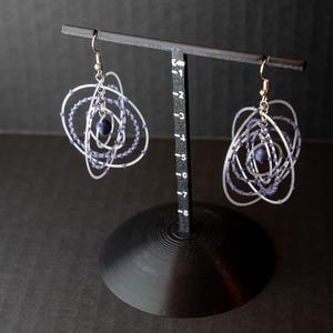 May include: A pair of silver-toned, orbital-shaped earrings with dark blue beads. The earrings are displayed on a black stand against a dark background. The design features multiple concentric circles, creating a unique, modern look.
