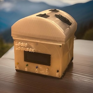 May include: A beige Logic Lockbox with a curved top and black latches. The front of the box has a small screen, buttons labeled A, B, C, and D, and the text "Logic Lockbox". The box is sitting on a wooden surface.