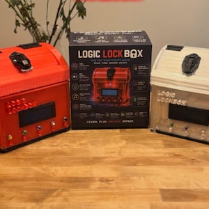 May include: Two Logic Lock Box puzzle chests, one red and one white, sit on either side of their retail packaging. Each chest features a black latch on the lid, a digital screen, and several small colourful buttons on the front panel.