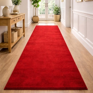 May include: A long, rectangular red rug in a hallway. The rug is a solid color and covers the majority of the floor space. The hallway has light-colored wooden floors and white walls. A wooden console table with a mirror and plants is visible.