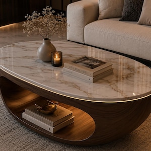May include: Oval-shaped coffee table with a marble top and a dark wood base. The table features a lower shelf and a warm, ambient light around the edge. Decorative items include books, a candle, and a vase with dried flowers.