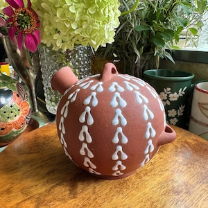 May include: A spherical terracotta vessel with a matte finish, featuring a small spout, a top handle, and a side handle. The surface is decorated with vertical rows of white, teardrop-shaped painted patterns arranged in a repeating design.